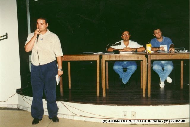 Dirigente fala ao microfone diante de mesa com outros participantes em auditório.
