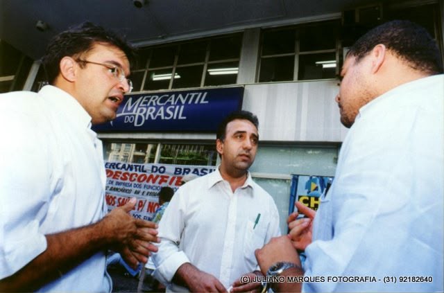 Três homens conversam diante de uma agência do Mercantil do Brasil em contexto de atividade sindical.