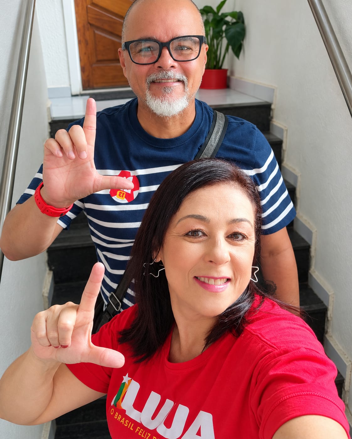 Juliano e Cynthia Marques juntos, sorrindo para a câmera e fazendo o gesto da letra L.