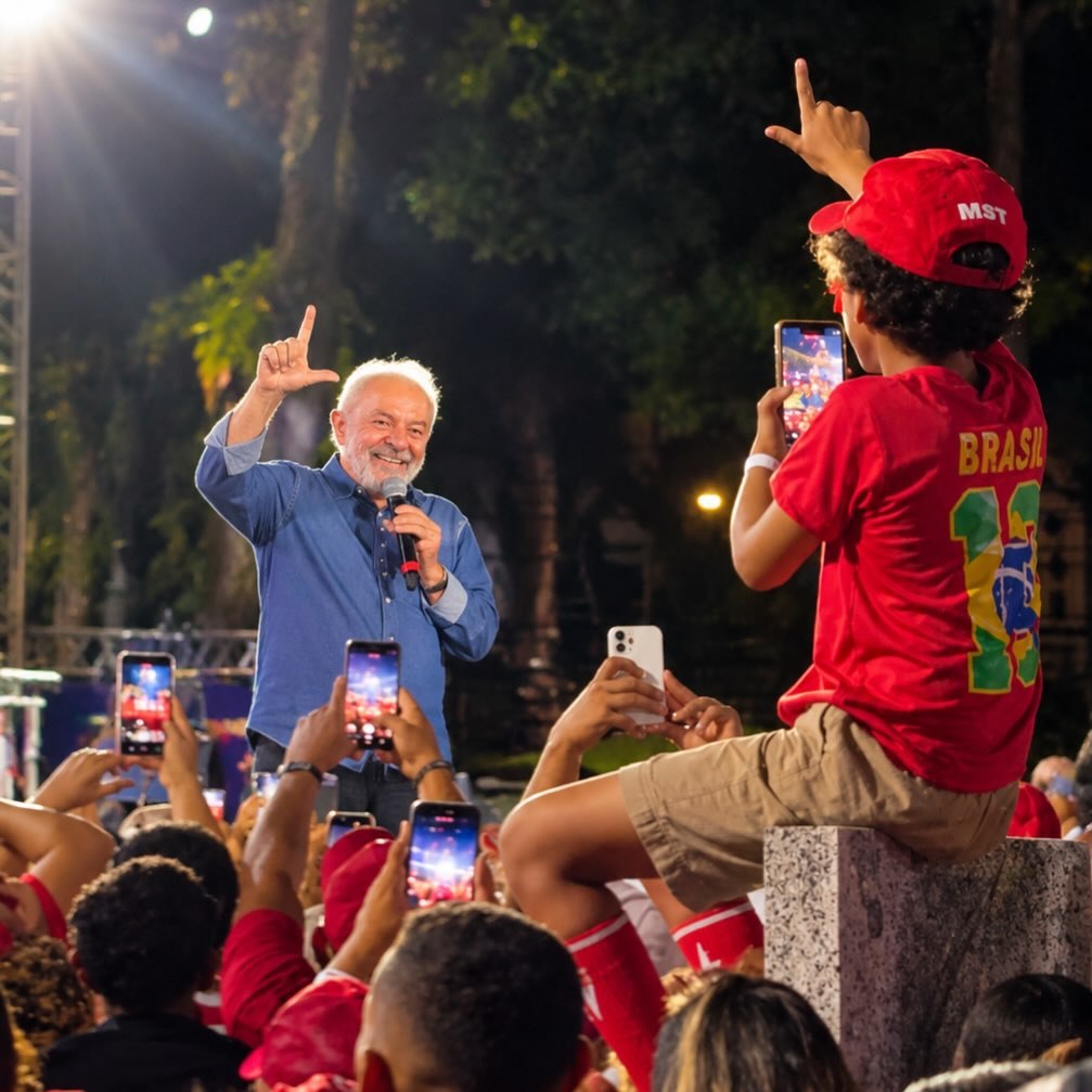 Registro de ato político com público fotografando uma liderança ao microfone